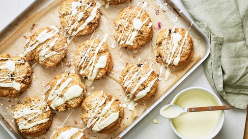 Blueberry and oat scones on a lined baking tray next to a bowl of melted white chocolate and pale green tea towel